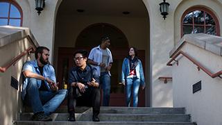 A group of students talking at the entryway to Cramer Hall. Two students are seated on the steps talking and two additional students are talking as they exit the doors.